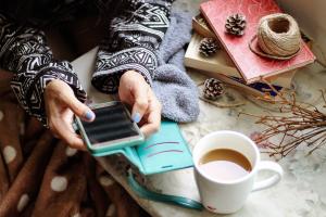 Person håller telefon i sina händer. Bilden tagen från ovan. En kopp kaffe och hantverksförnödenheter på bordet bredvid händerna och telefonen.